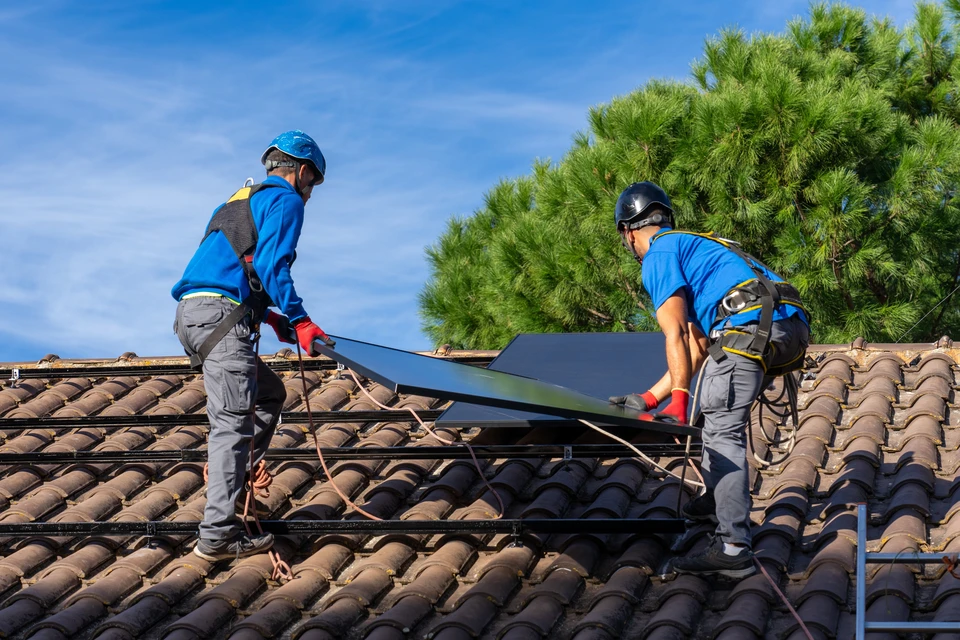 Technicien en combinaison verte installant ou entretenant des panneaux solaires photovolta&iuml;ques sur toiture, portant gants de protection, utilisant outil de serrage sur syst&egrave;me de fixation