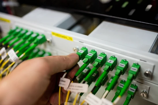 Intérieur d'un centre de données, technicien en pull bordeaux manipulant des câbles de fibre optique jaunes dans une baie de brassage réseau